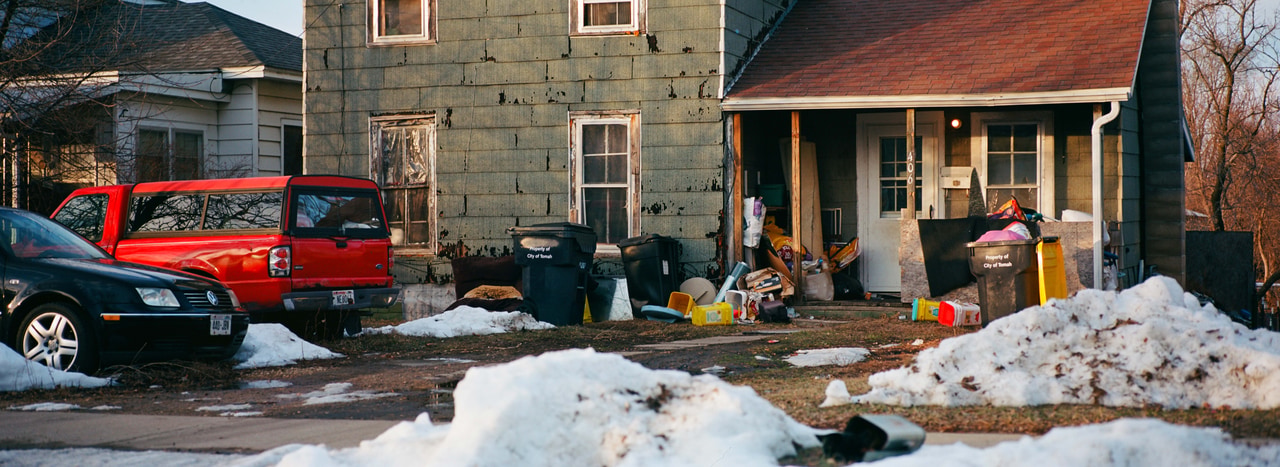 A red vehicle beside a weathered house and banks of melting snow